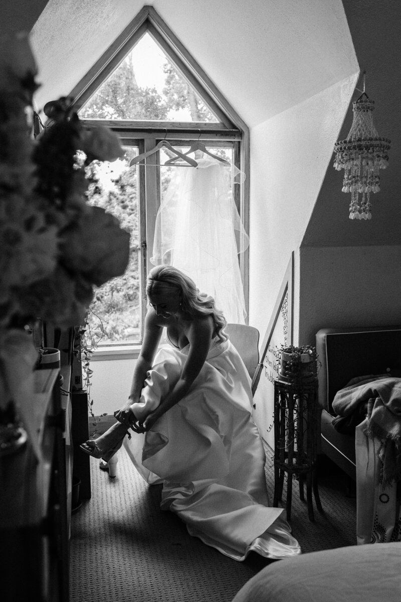 black and white photo of bride in front of window putting shoes on in waimumu gore
