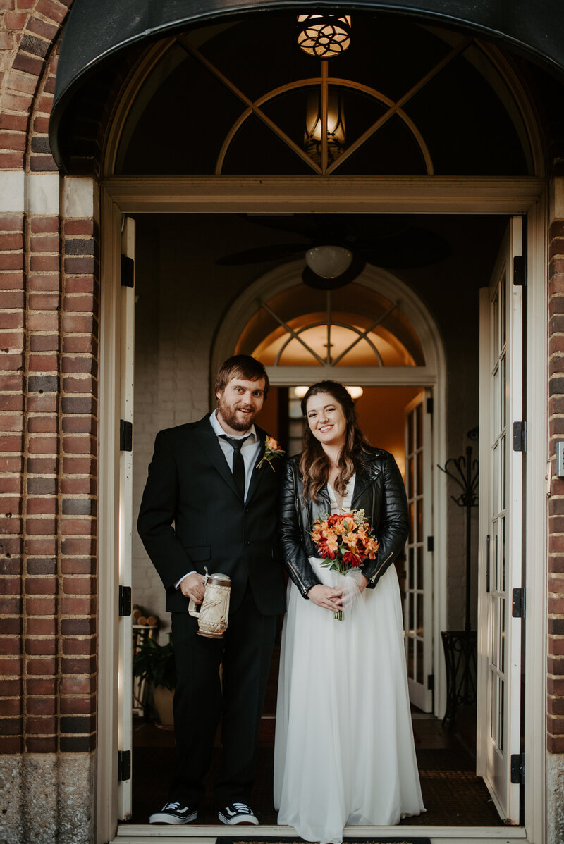 couple standing under historic doorway bride wearing black leather jacket holding red yellow flowers