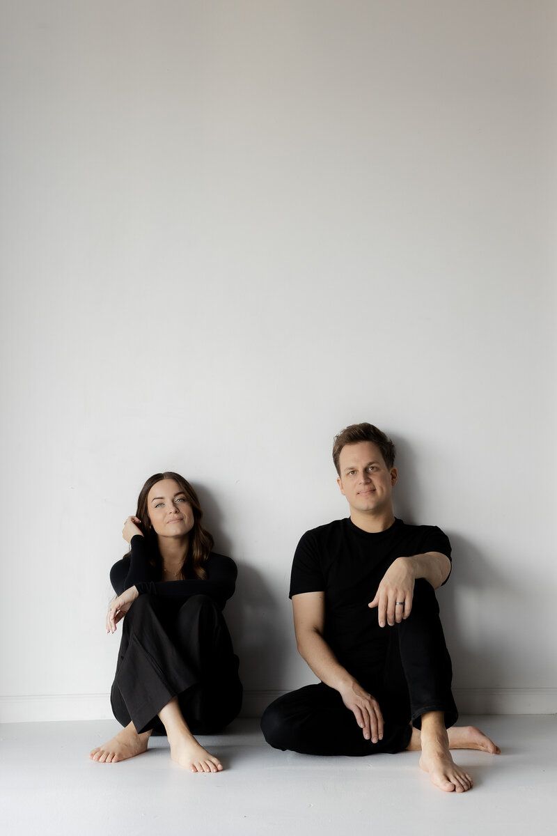 Todd and Madi sitting barefoot in a clean, minimal studio, showcasing their style as TM Photo Co, a wedding and lifestyle photography team.