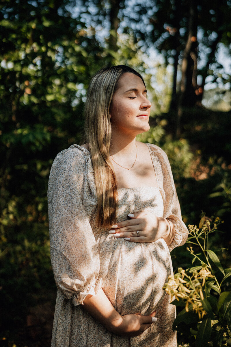Maternity portrait of a mother facing the sunlight holding her belly with flowers and the shadows of flowers on her face, body and pregnant belly. 