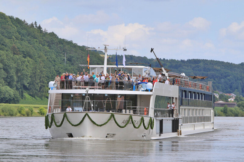 A large river cruise ship travels along a calm waterway, decorated with green garlands along the bow. Passengers stand on the top deck enjoying the view, surrounded by lush green hills and a partly cloudy sky.