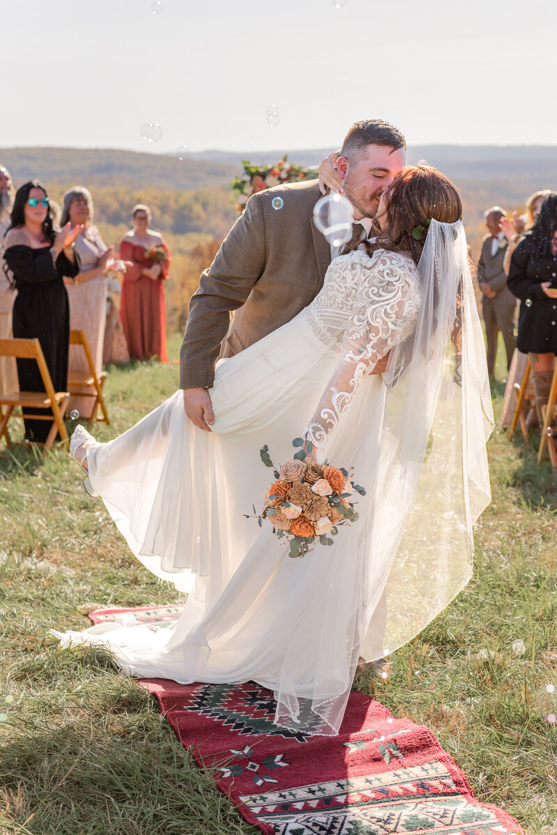 Newly married couple shares a joyful kiss during their outdoor fall wedding in southwestern Pennsylvania. Surrounded by guests and golden foliage, the groom dips the bride at the ceremony’s end, with bubbles floating through the air.