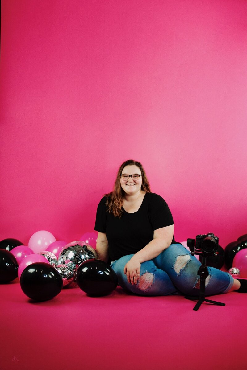portrait of mara shields, owner of M20 Media, sitting and smiling, with her camera, gimbal, balloons, and a hot pink background