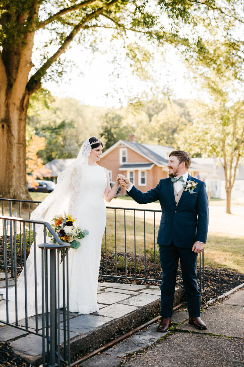 Richmond weddings with Virginia wedding photographer capturing bride and groom walking down a stone path together as the groom helps his bride over a step in the path with colonial buildings in the distance and a large tree shadowing over them