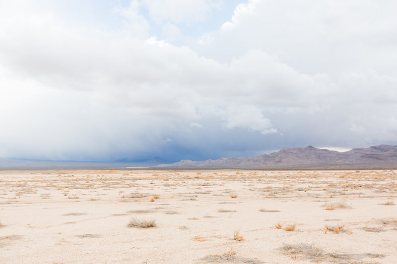 A vast desert landscape with dry grass patches and distant mountains beneath stormy clouds outside of Las Vegas