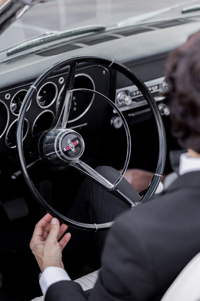 A groom in a sleek black tuxedo grips the wheel of a vintage Oldsmobile, capturing the essence of classic luxury and timeless elegance. This close-up shot of the retro dashboard and polished chrome details evokes the nostalgia of a stylish getaway in Palm Springs, California.