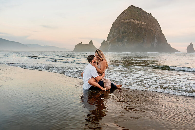 a photo of a couple in cannon beach sitting together in  the water with haystack rock in the background 