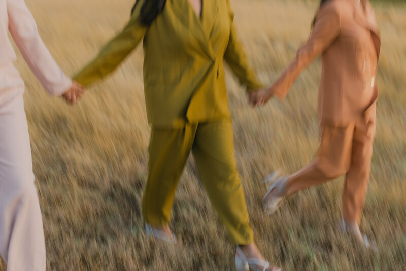 Three women walk hand-in-hand through a golden field wearing pastel and earth-tone suits, captured in a dreamy, motion-blur editorial style. Greenville South Carolina portrait photographer – Dani Lacey Photographs.