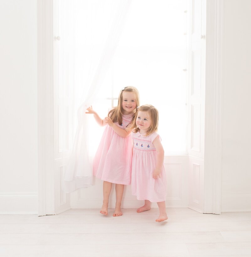 Two young sisters stand together in front of a bright window at Dandelion Studio in Moira, Northern Ireland. Both girls are dressed in matching pink dresses with delicate embroidery. The older sister is holding the sheer curtain open while the younger sister stands close, both smiling softly. The scene captures a gentle and innocent moment, highlighting their close bond and playful curiosity.