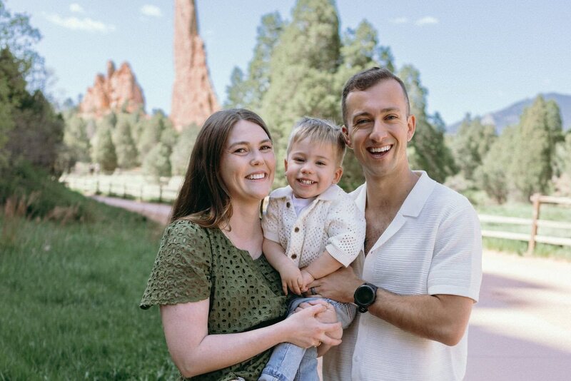 A smiling mom and dad with their toddler in front of red rock formations at Garden of the Gods.