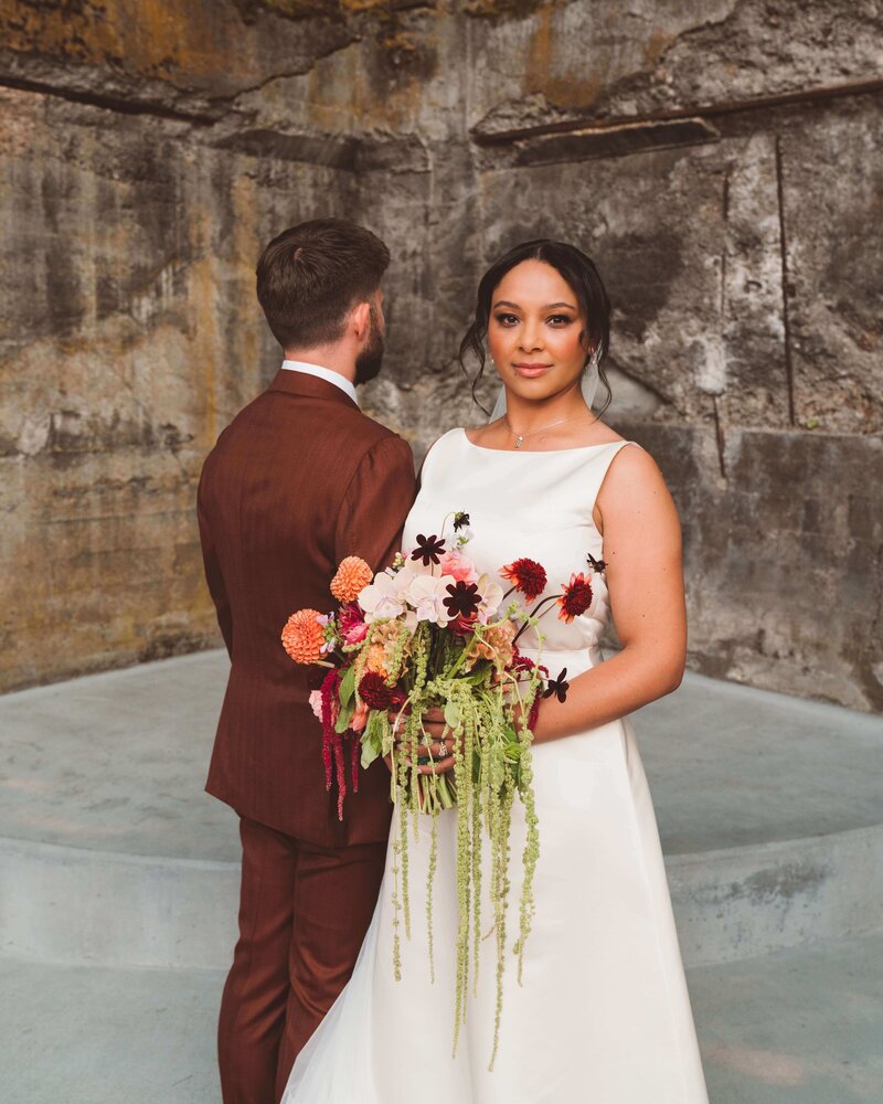 Bride looks at camera holding a colorful and bold wedding bouquet. The bouquet is wild and cascading with green amaranth. The bride is wearing an all-white, modern wedding dress. The groom is on the left and is facing away from the camera. The groom is wearing a warm brown suit. The background is  rough, textured stone walls and a concrete platform. Location is The Ruins wedding venue in Hood River, Oregon.