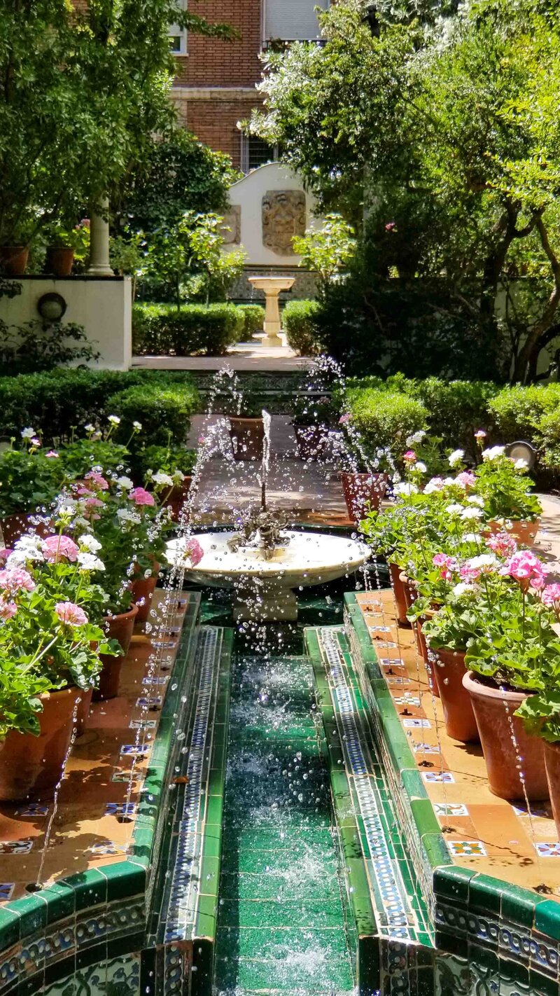 Ornate Moorish-style courtyard garden at the Museo Sorolla in Madrid, Spain, featuring a green-tiled water channel with fountains, surrounded by terracotta planters filled with pink and white flowers, and a stone fountain in the background ©Stephanie Dosch | theViatrix Luxury Travel