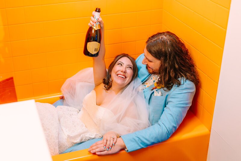 A wedding couple sitting in a retro bathtub in their wedding attire holding a bottle of champagne smiling at each other