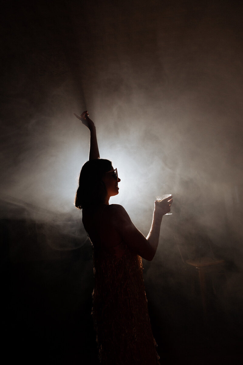 The silhouette of a person with one arm up and the other holding a drink 