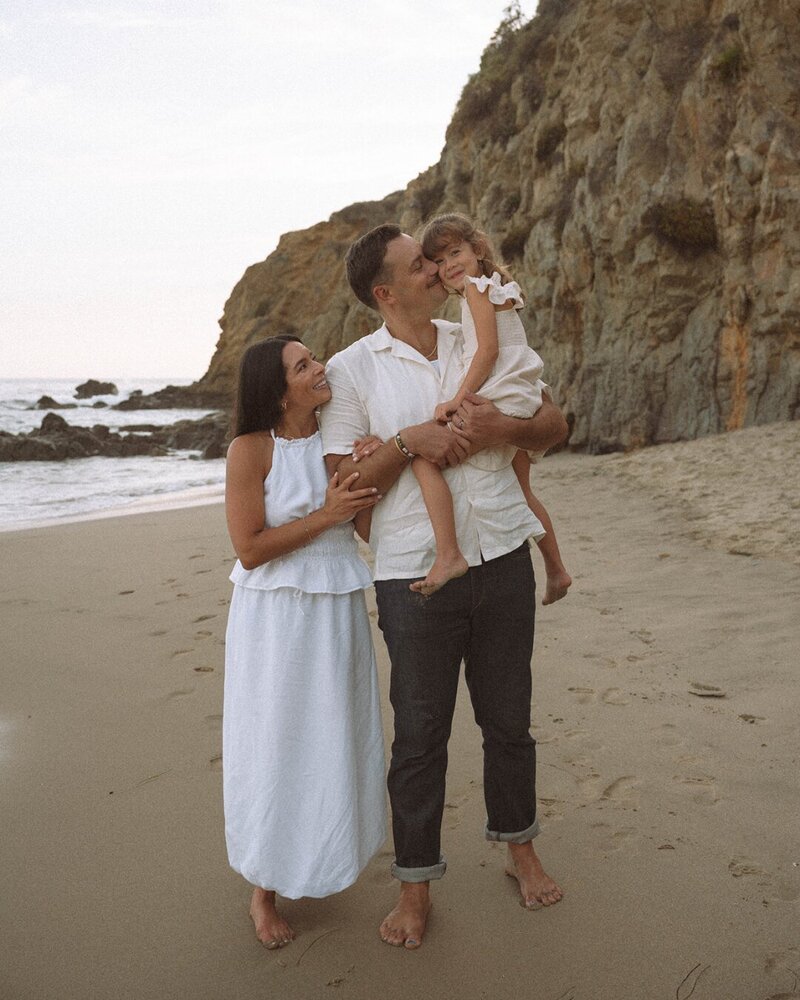 Family standing barefoot on the sand at Laguna Beach, with rocky cliffs in the background during sunset.
