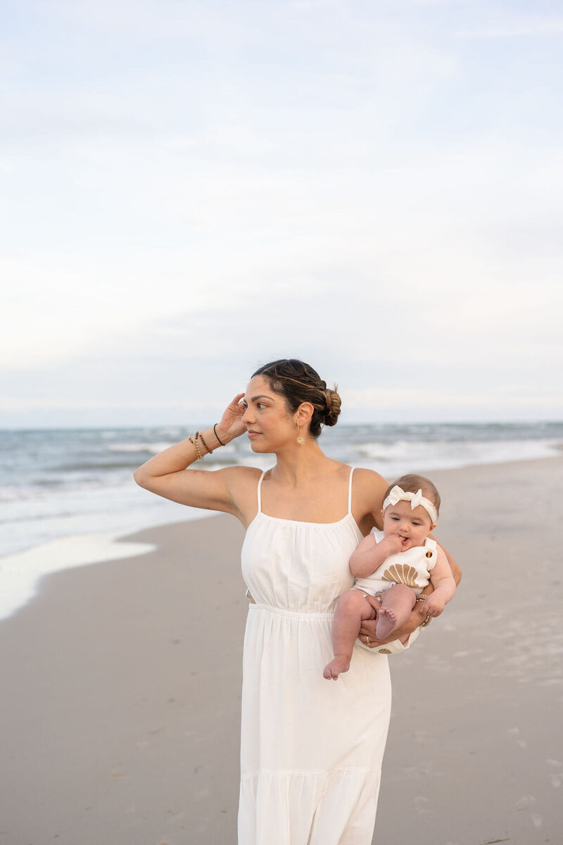 mother wearing a long white dress holding baby girl as she gazes over the ocean  sweeping a strand of hair behind her ear