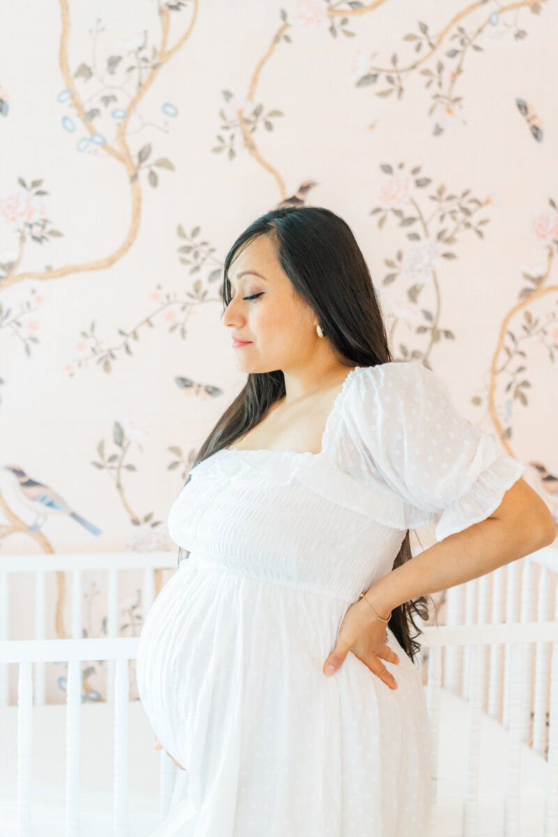 Austin Maternity Photographer | A pregnant mother in a long white dress with long dark hair stands in front of a white crib in a pink nursery. 