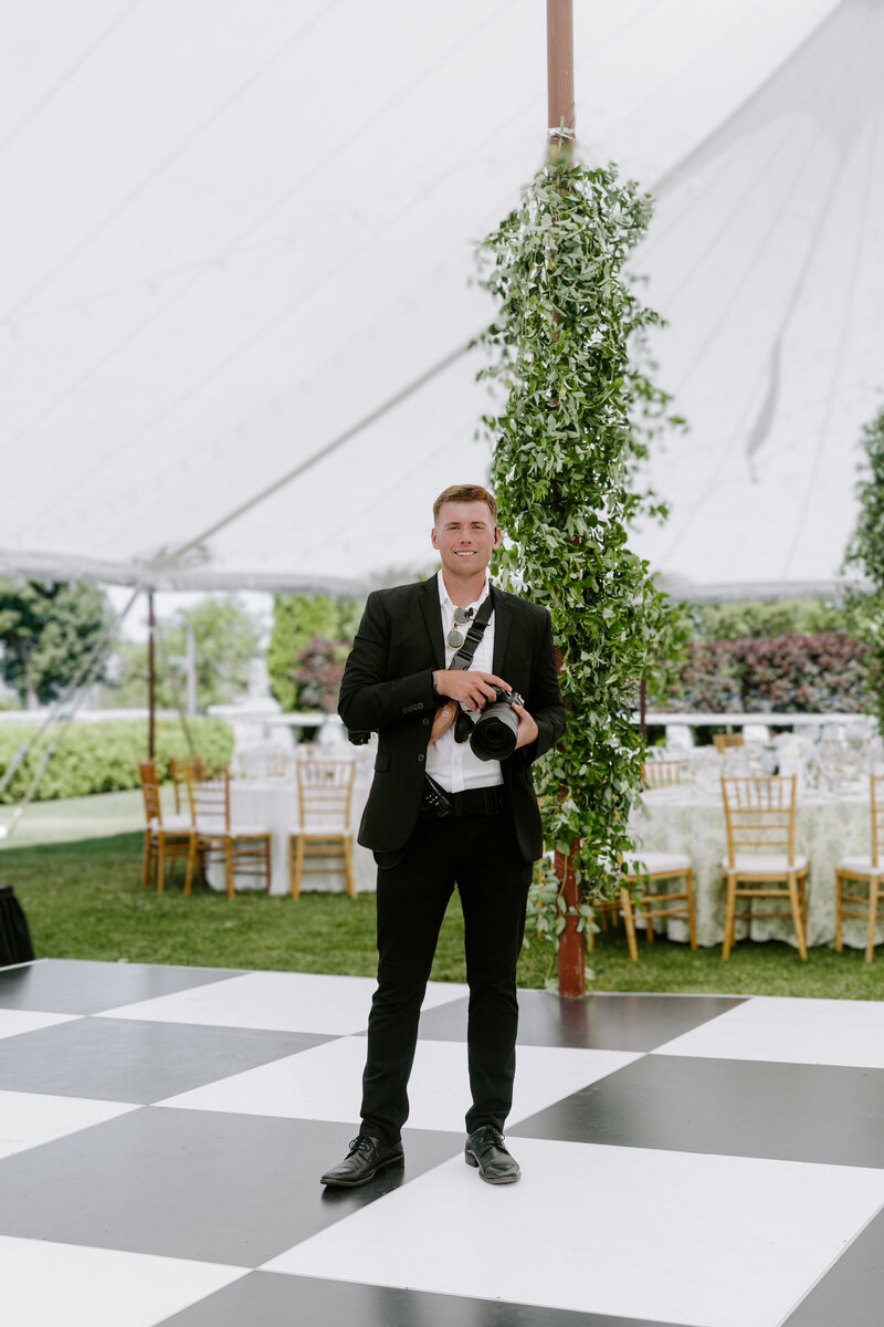 Fine art wedding photography Maine wearing a black suit while holding his camera and smiling