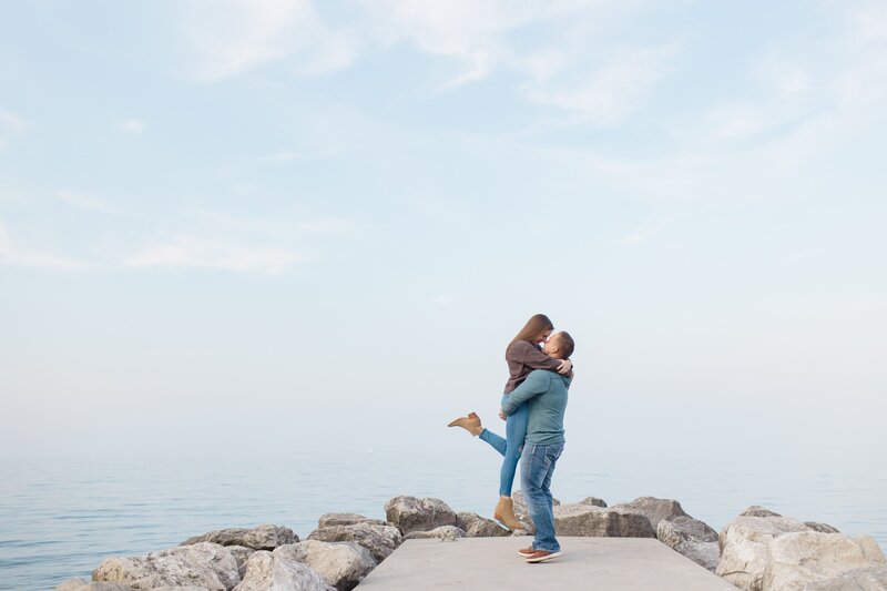 A man picking up his fiance and they're face to face smiling at each other on a pier in edgewater beach near cleveland ohio