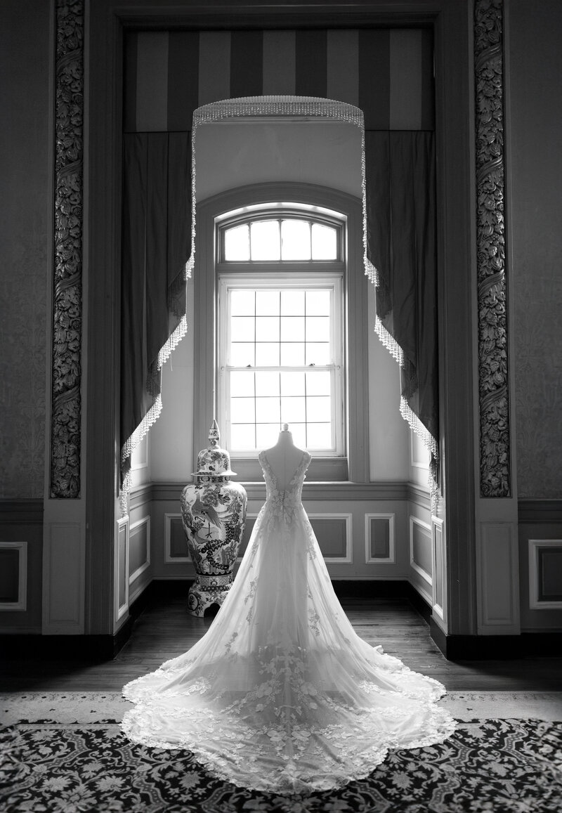 A bridal gown displayed on a mannequin stands elegantly in front of a large window with draped curtains, letting in soft light. The intricate details of the dress are highlighted, with a decorative vase nearby. Black and white color scheme.