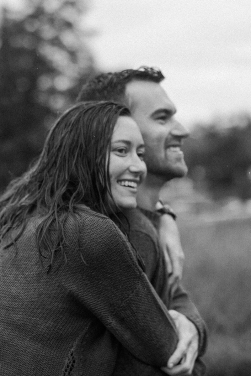 A couple embraces from behind, surrounded by a natural setting, conveying intimacy and connection in a black-and-white photograph.