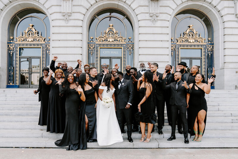newlyweds kiss surrounded by wedding party in front of city hall