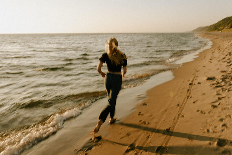 A senior girl in relaxed attire runs along the shores of Lake Michigan, illuminated by the golden hues of a breathtaking sunset, captured by a Michigan senior photographer known for crafting natural and emotive portraits.