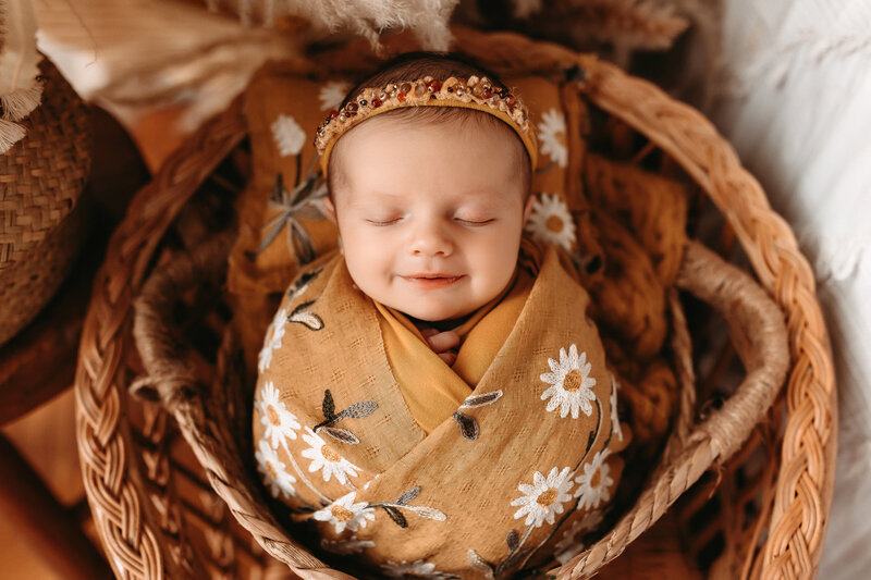 Newborn baby girl smiling while wrapped in a mustard floral swaddle.