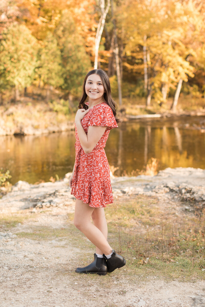 Senior girl standing by a river smiling at the camera with beautiful fall colors in the background