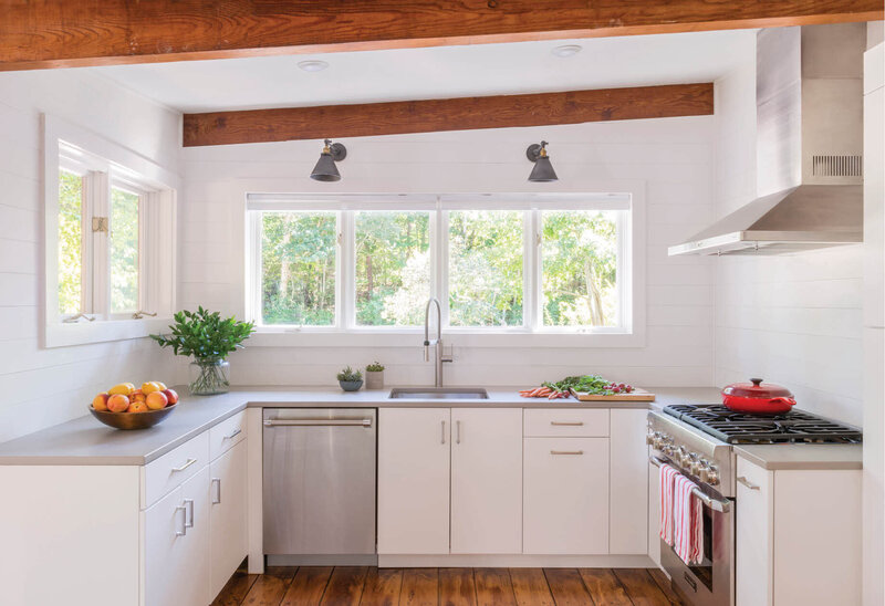 Cape Cod interior designer creates a Falmouth kitchen with white cabinets, reclaimed wood beams and posts, white shiplap walls, and new sconces above the windows.