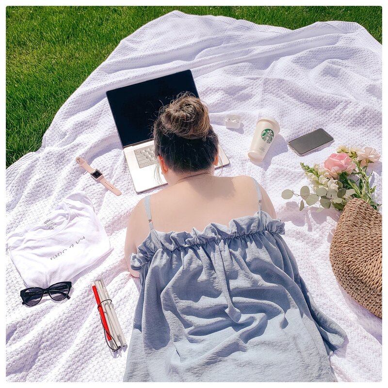 On a white blanket outdoors on the green grass, Anne, an Asian woman, is on her stomach working on her laptop computer. In a clockwise description items surrounding her are her white cane, sunglasses, Braille beaded T-shirt that reads “purpose in view”, Apple Watch, Apple AirPods, Starbucks cup, iPhone, and florals in a straw purse. She is wearing a blue tank top with spaghetti straps. The tank has a delicate feminine ruffle.]