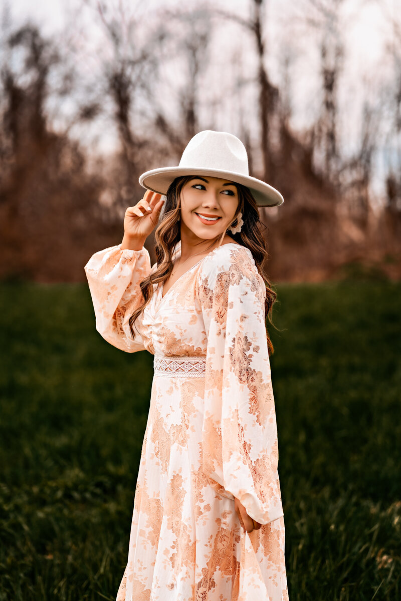 Emily Belanger an elopement photographer in a light pink dress and white hat looks over her shoulder smiling while stand in a green field with the woods blurred in the background