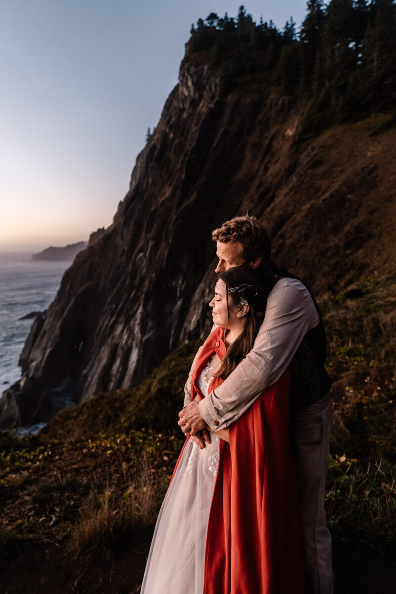 During the twilight of their Oregon elopement, a couple in their wedding attire snuggles on a coastal cliff .