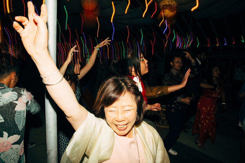 The bride's mom joyfully dances along to the music at the wedding reception with other guests dancing in the background under the colorful lights in Dos Pueblos Orchid Farm.