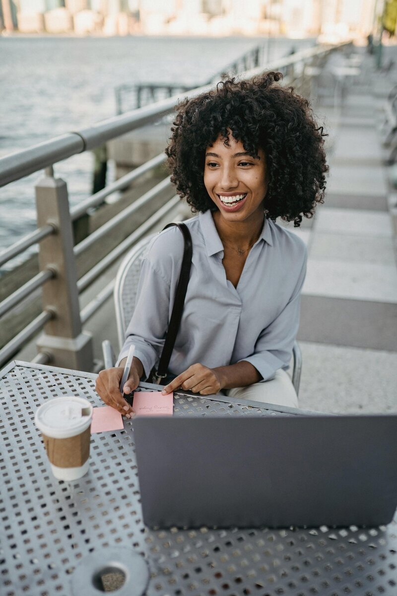 woman working on a laptop outside by the water with a coffee and post it notes
