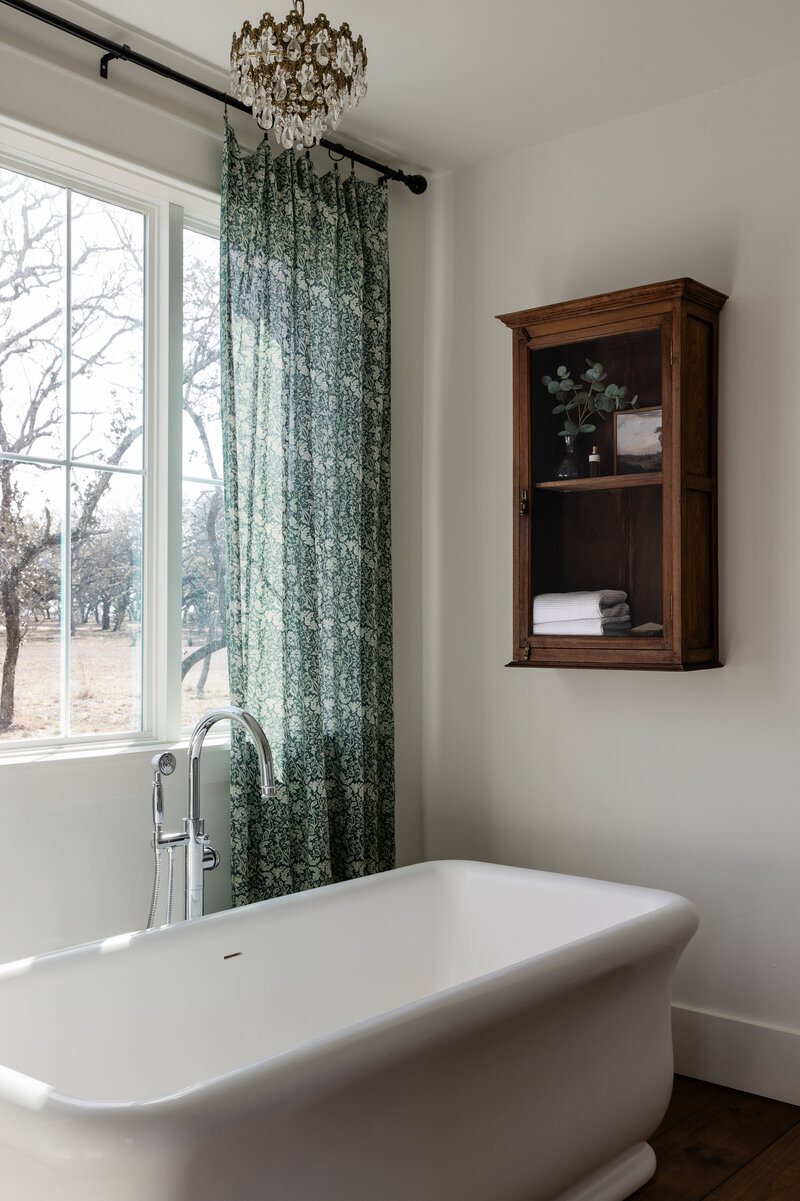 A massive soaking tub, vintage storage cabinet, and floral drapes bring this primary bathroom to life in the Texas Hill Country. Designed by Jessa Myer Interiors.