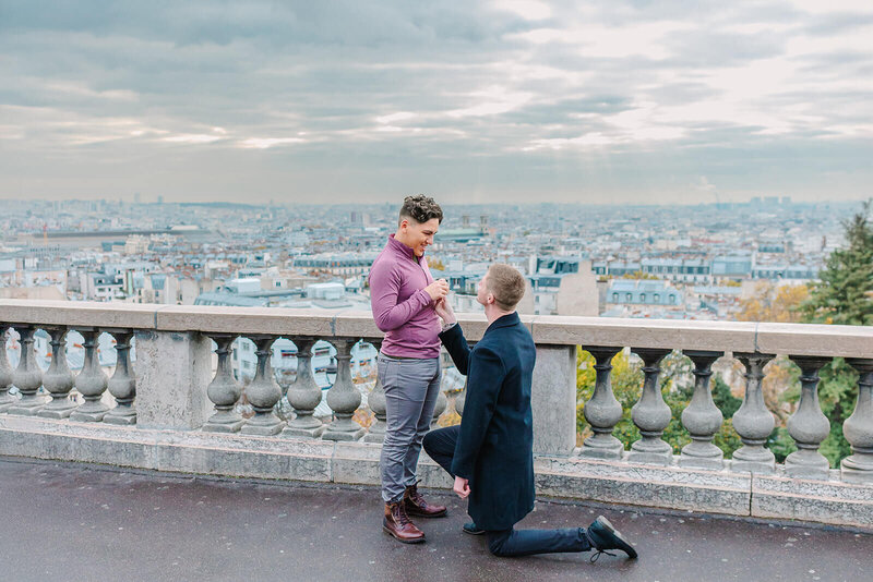Gay couple during a Paris proposal at Sacré-Cœur in Montmartre — one man kneeling as the other joyfully accepts.