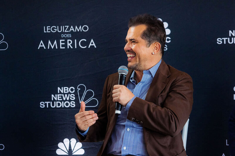 John Leguizamo speaks into a microphone at an event