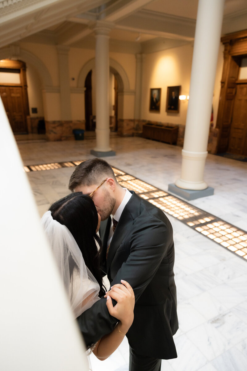 Bride and groom sharing an intimate kiss inside a marble courthouse, framed by white columns and soft indoor lighting.