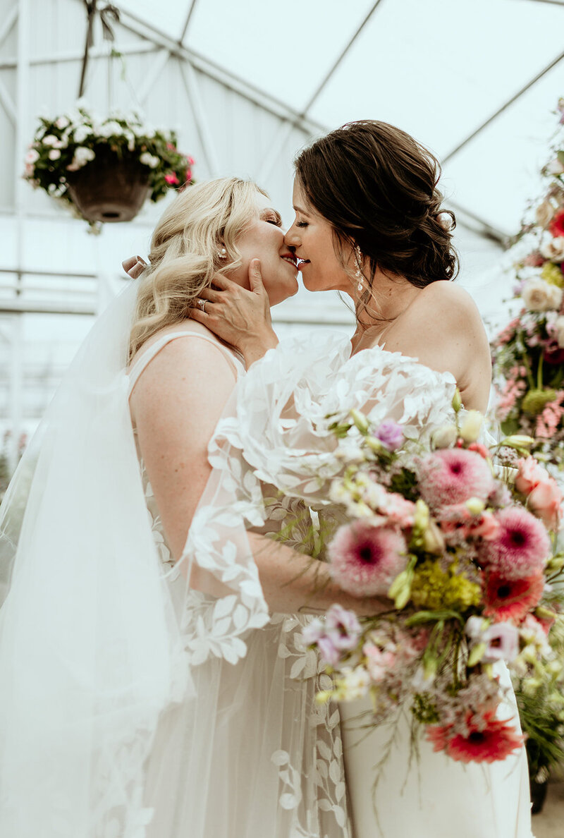 wife and wife holding each other and kissing in gorgeous gowns in beautiful greenhouse