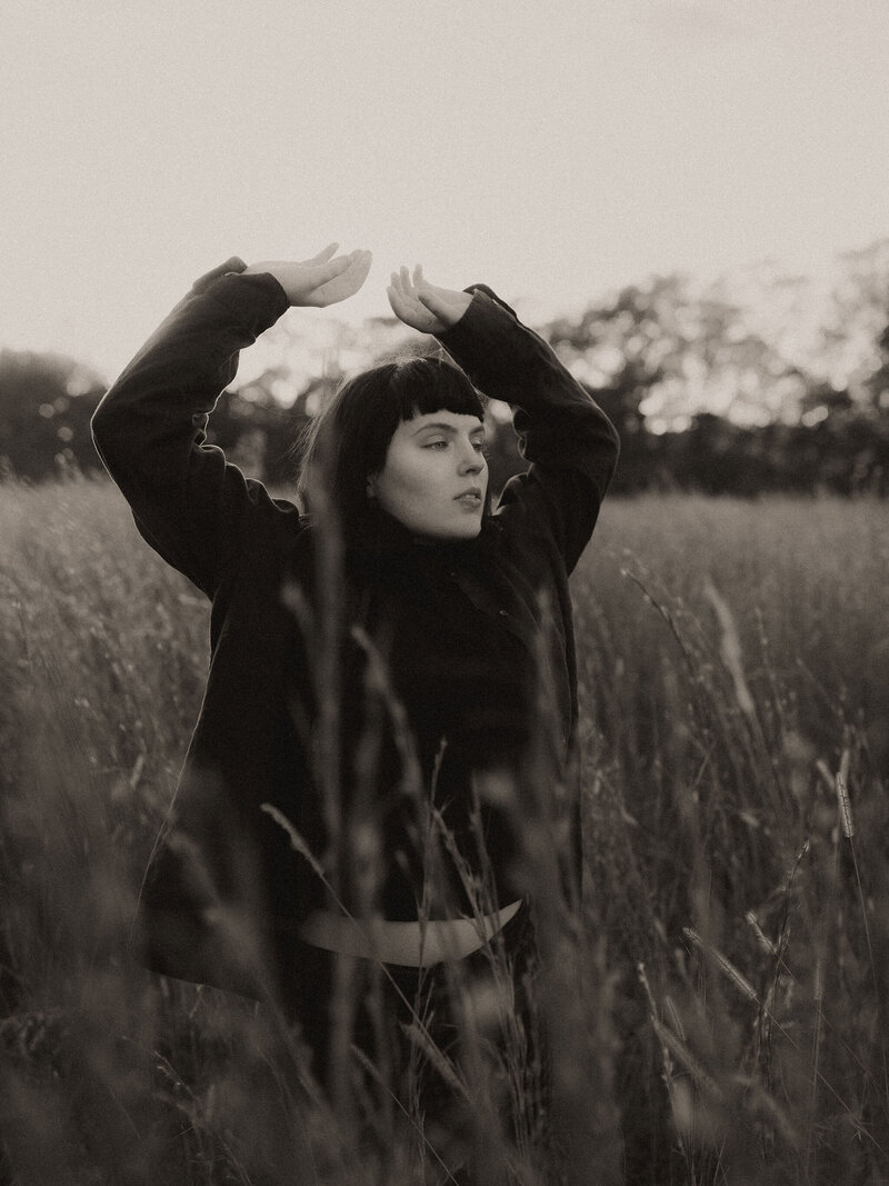 A woman with sharp bangs and a black turtle neck posing in an overgrown field. 