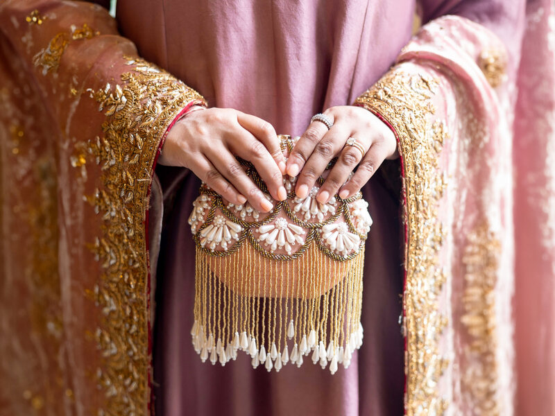 A woman in a pink and gold embroidered outfit holds an ornate clutch. The clutch is gold with pearl embellishments and fringe details. The womans hands are adorned with rings.