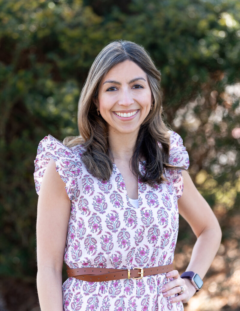A New Jersey female psychologist specialize in trauma, anxiety, & depression in a floral dress, hand on her waist, and smiling at a park.