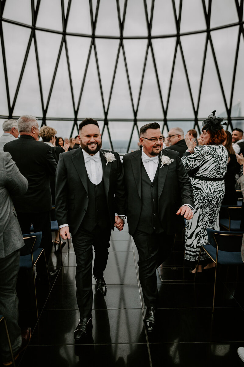 A same sex couple wearing black tie suites walk up the aisle at their wedding at The Gherkin in London. 