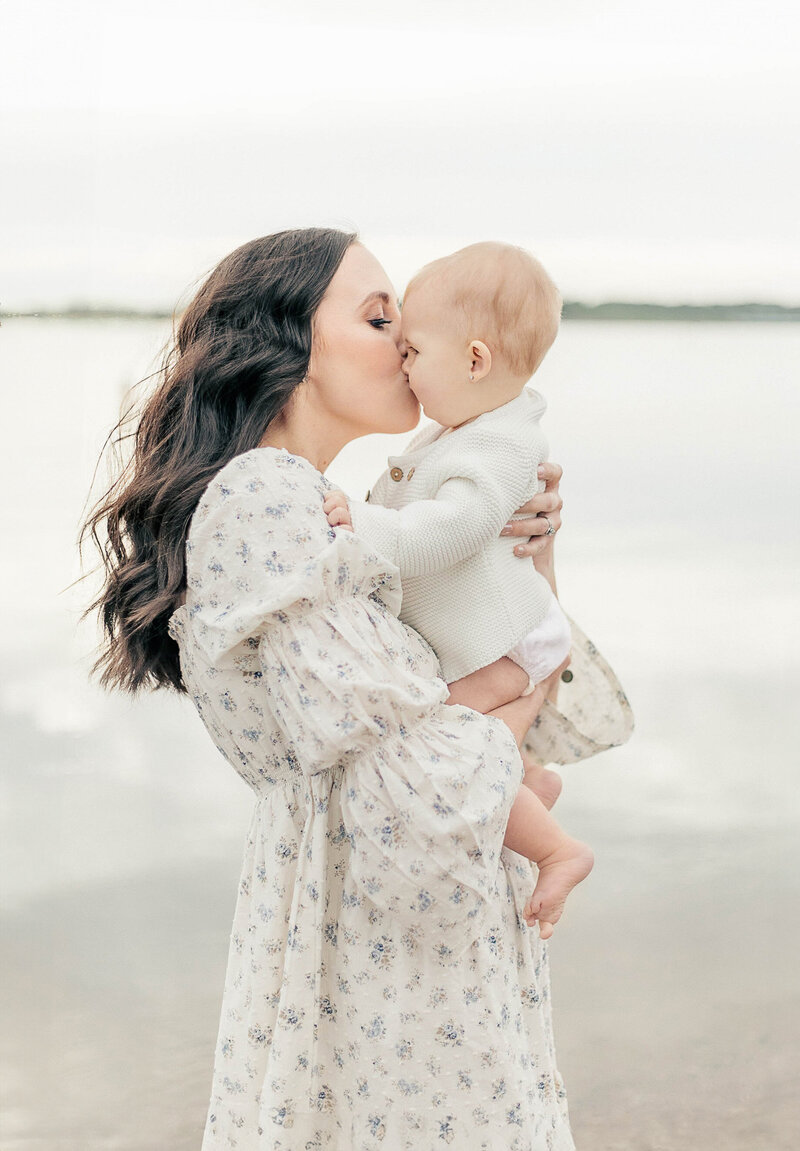 Mother in a floral dress lovingly kisses her baby during a lakeside family photography session in Kansas City.