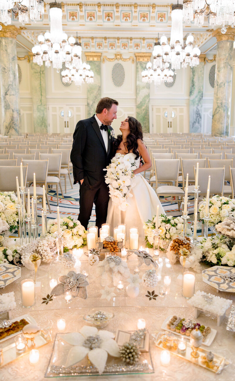 A Persian bride and groom smile at each other in an elegant wedding venue with chandeliers. Candles, flowers, and crystals surround them on the sofreh table, creating a romantic atmosphere.