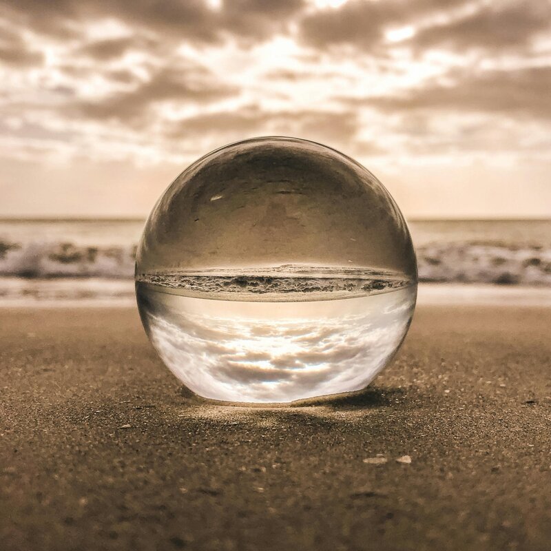 Image of a glass globe in the sand at the beach that is reflecting the sun and clouds