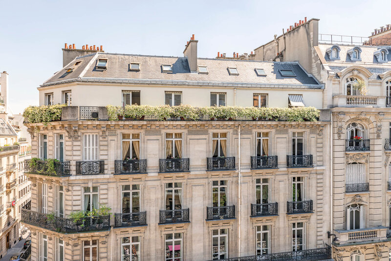 Elegant Parisian-style apartment building with wrought-iron balconies, tall windows with curtains, and greenery along the rooftop terrace under a clear blue sky.