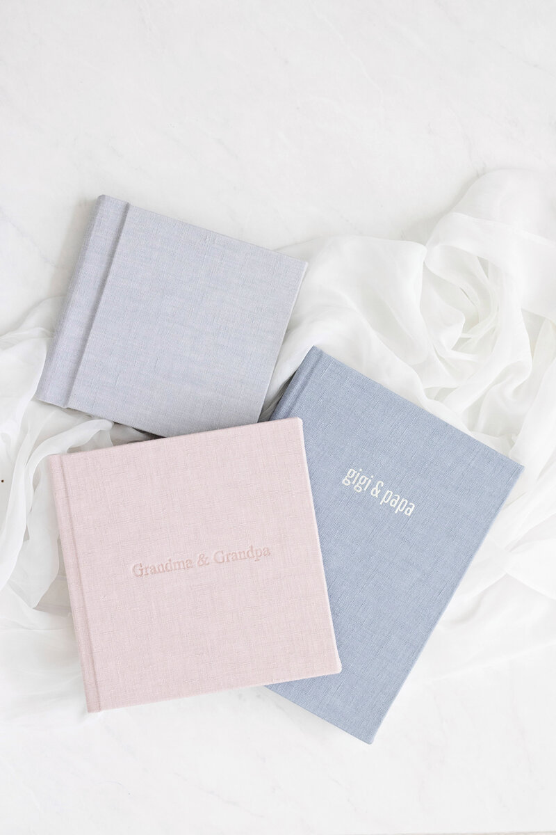 Three photo albums (gray, blue, and light pink), laid on top of one another on top of a white cloth background.