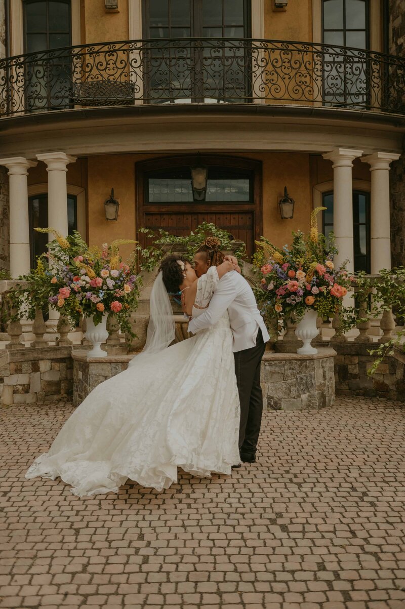 Newlyweds share a romantic kiss as the groom dips the bride in front of a grand, castle-like venue with arched windows, ornate railings, and stone columns. The bride's voluminous gown and veil cascade to the cobblestone courtyard, framed by vibrant floral arrangements in tall white urns.